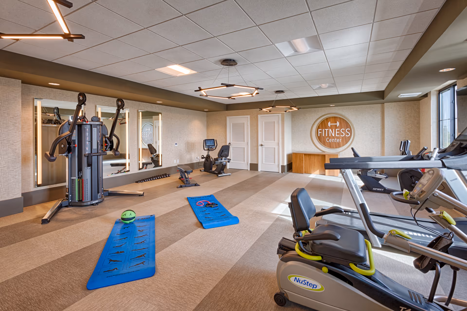 A bright and spacious fitness center with exercise equipment including a NuStep recumbent bike, treadmills, a multi-functional weight machine, and two blue exercise mats on the floor. The room has beige walls, large mirrors, modern ceiling lights, and a wooden sign on the wall that reads 'Fitness Center'.