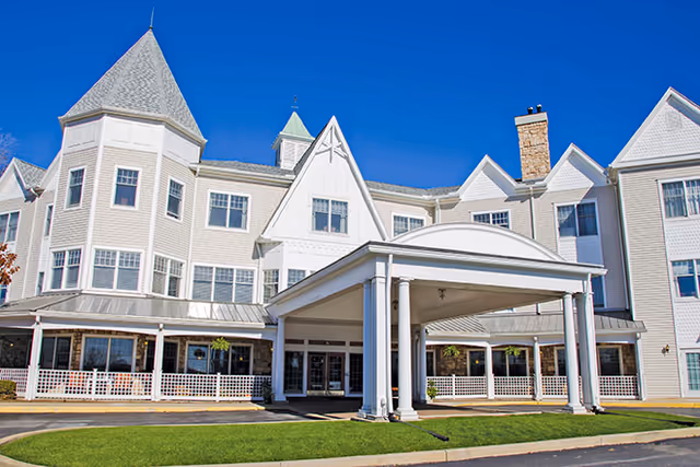 White multi-story senior living building with a covered porte-cochere entrance and turrets under a clear blue sky.