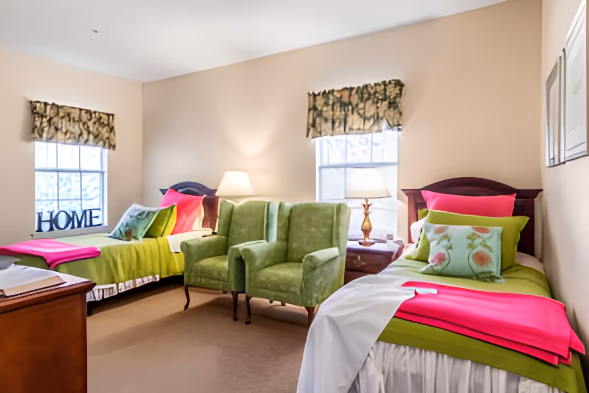 A bright bedroom with two twin beds dressed in green and pink bedding, two green armchairs between them, bedside lamps, and windows with valances.