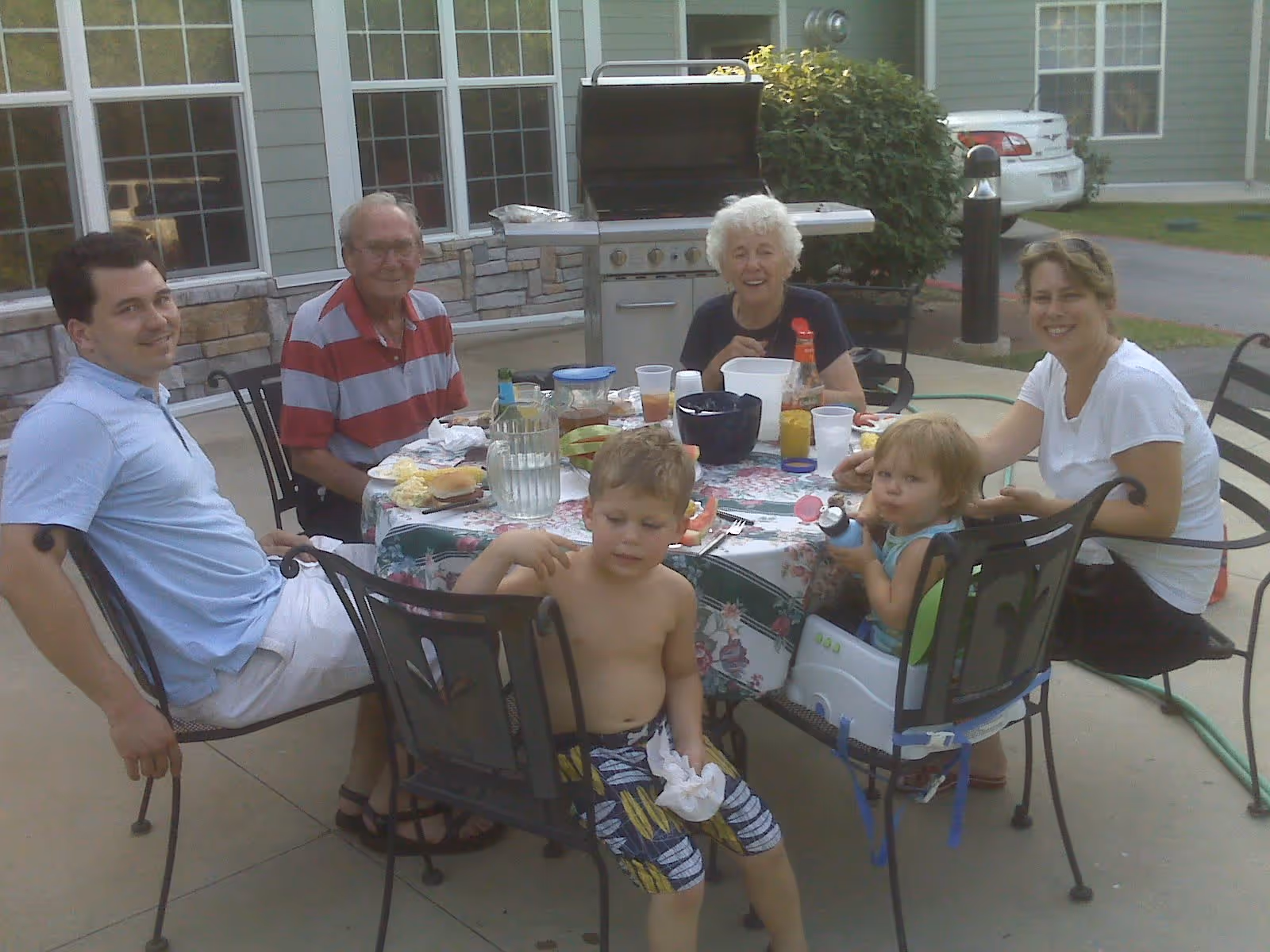 A group of six people, including two children and four adults, sitting around a round outdoor table with food and drinks. They are on a patio outside a building with large windows and a grill in the background. The scene appears to be a casual outdoor meal or gathering.
