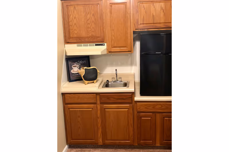 Small kitchenette with wooden cabinets, a compact sink, a black mini-fridge and countertop decor including a coffee sign and small chalkboard figure.