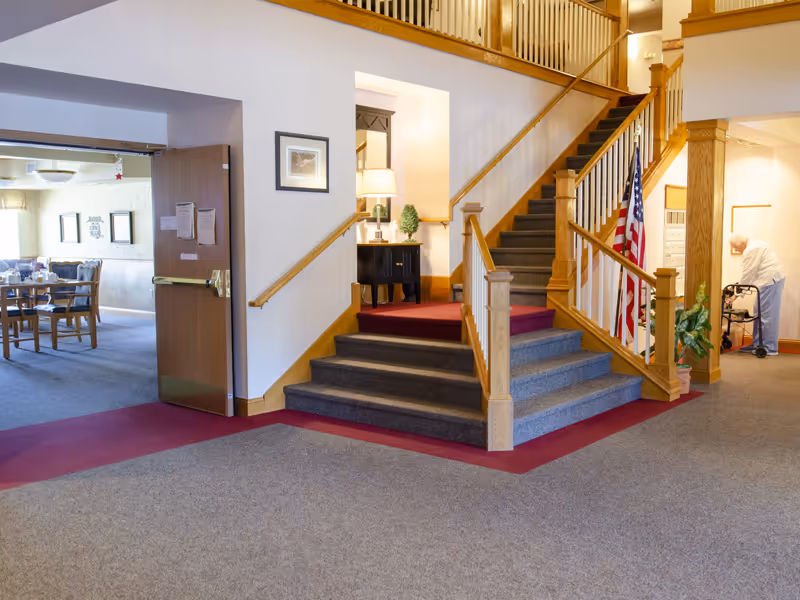 Interior view of a senior living facility showing a carpeted staircase with wooden railings, an American flag, a small table with a lamp and plant, and an elderly person using a walker near a wall with mailboxes. To the left, there is an open door leading to a dining area with tables and chairs.