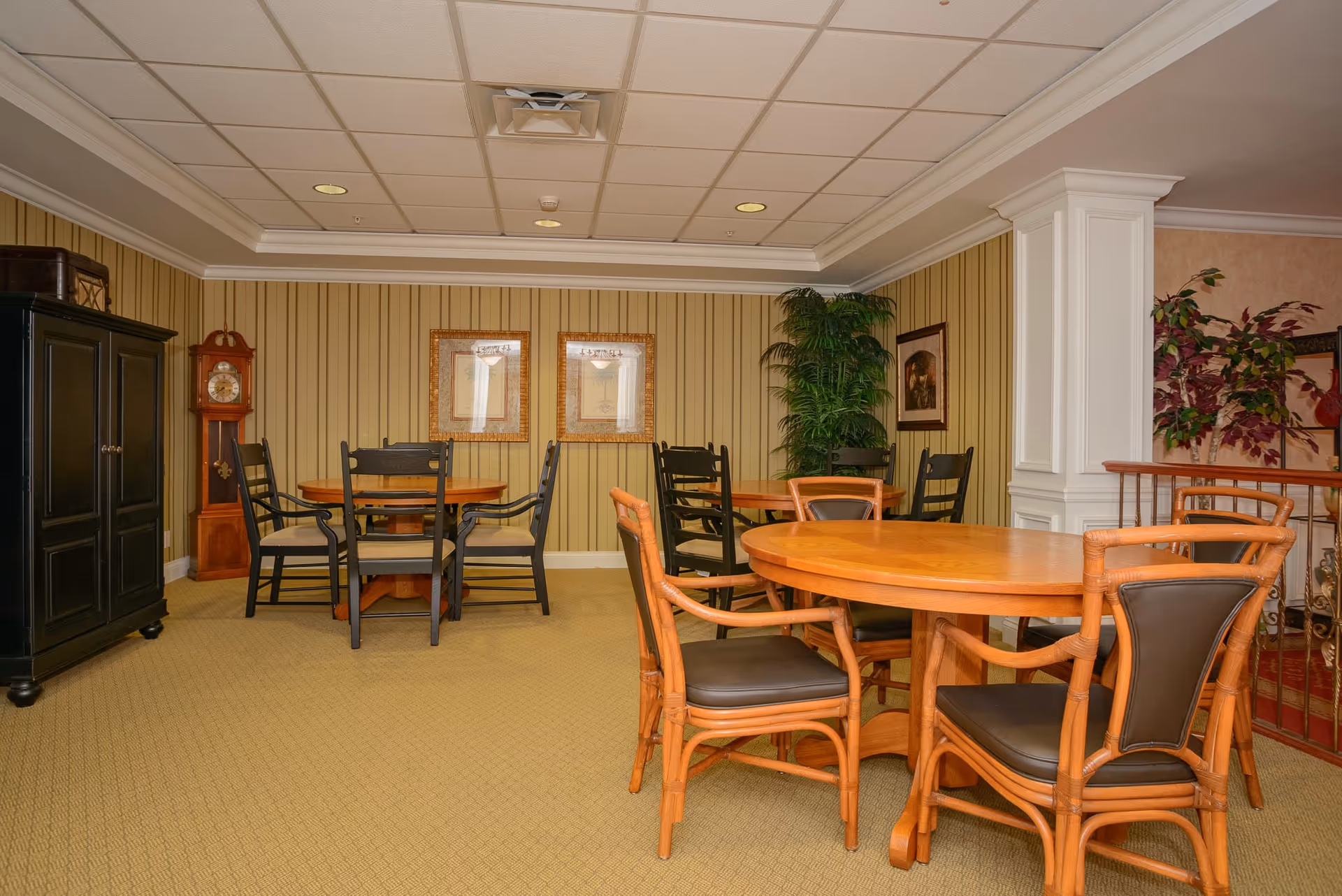 Interior view of a common area in a senior living facility featuring multiple round wooden tables with chairs around them. The room has beige carpet, striped wallpaper, framed artwork on the walls, a tall grandfather clock, a black cabinet, and several potted plants. The ceiling has recessed lighting and a ventilation system.