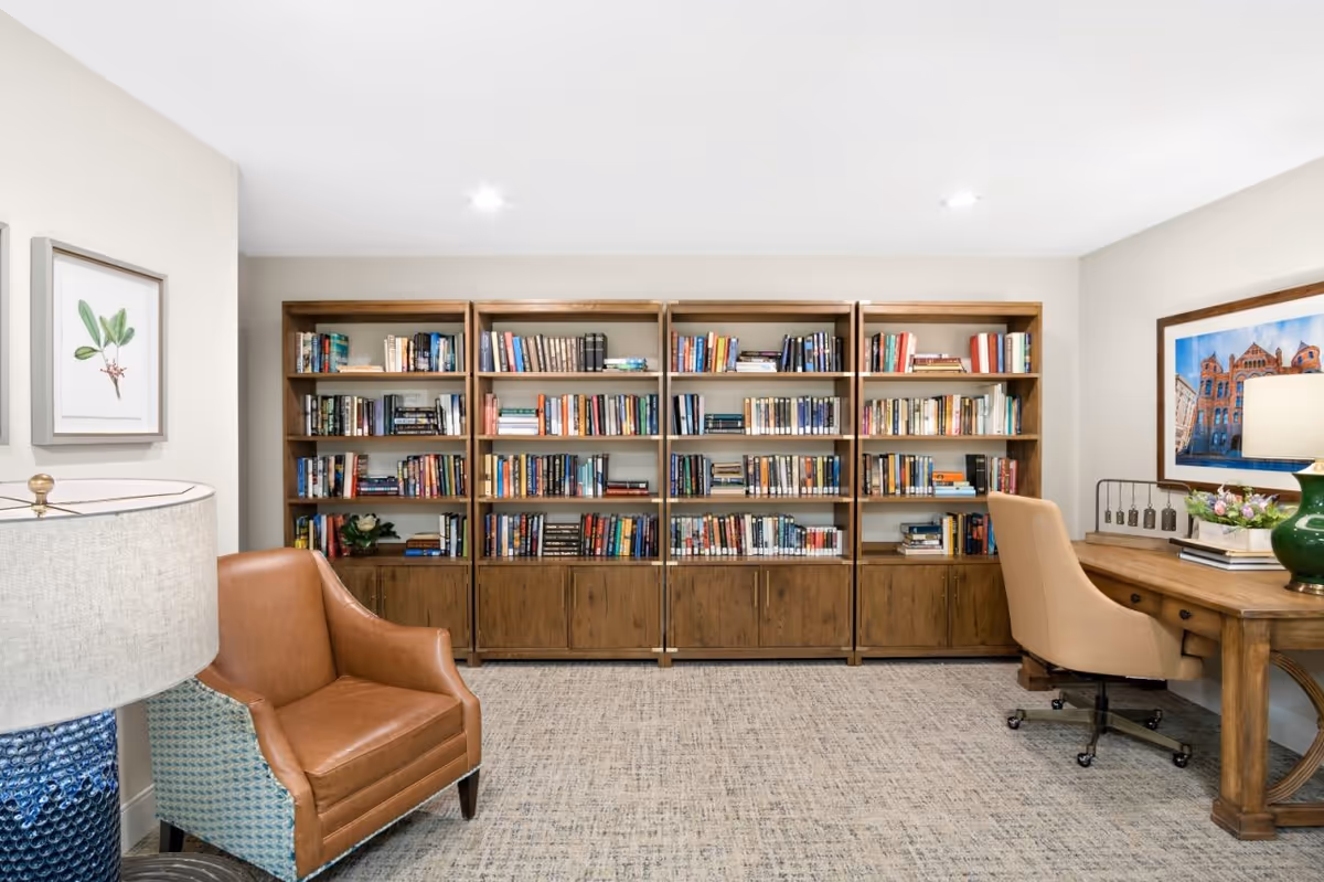 A cozy reading and work area featuring a large wooden bookshelf filled with books against the back wall, a brown leather armchair on the left, and a wooden desk with a beige office chair on the right. The desk has a green lamp, a framed picture on the wall above it, and some decorative items. The room has light-colored walls and carpeted flooring.