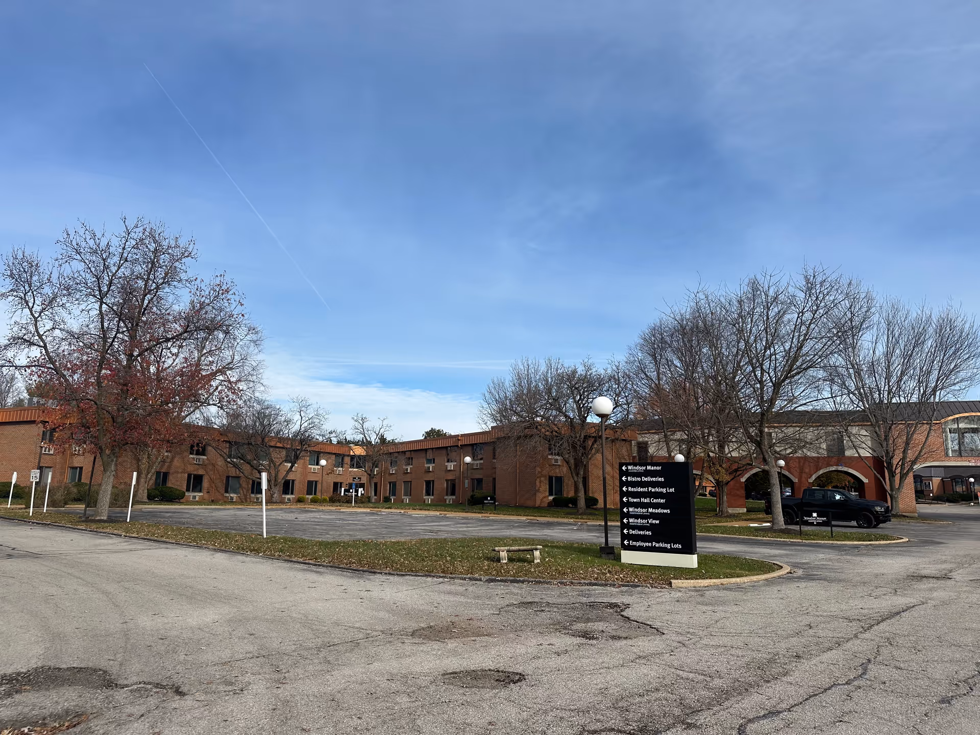Exterior view of a senior living facility named Windsor Estates of St. Charles, showing a large brick building with two stories, leafless trees, a parking lot with a few cars, and a signpost with directional information for various parts of the facility under a clear blue sky.