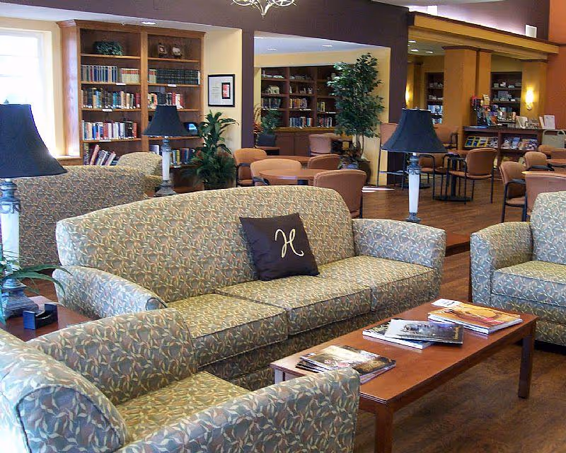 A cozy living room area in a senior living facility featuring patterned sofas and armchairs arranged around a wooden coffee table with magazines. The background shows bookshelves filled with books, several tables and chairs, potted plants, and warm lighting.