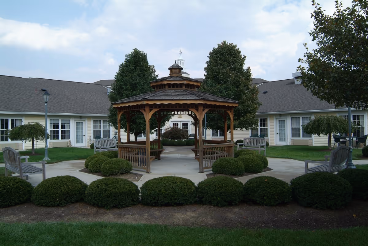 Outdoor courtyard area at Spring Hills Hunters Creek featuring a wooden gazebo surrounded by neatly trimmed bushes and benches. The courtyard is enclosed by a single-story building with multiple windows and doors, and there are several trees and lamp posts around the area under a partly cloudy sky.