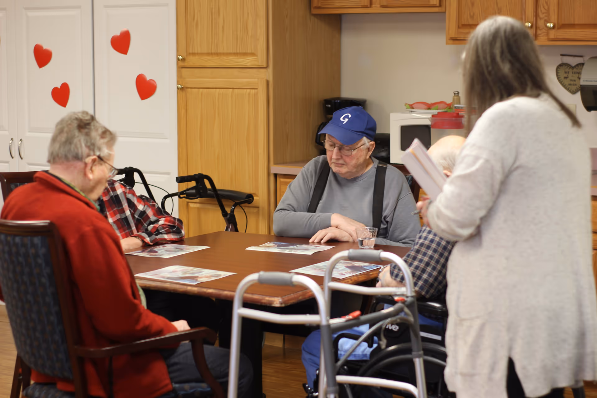 Several elderly residents sit around a table in a communal room while a staff member stands nearby, with walkers visible and heart decorations on a door.