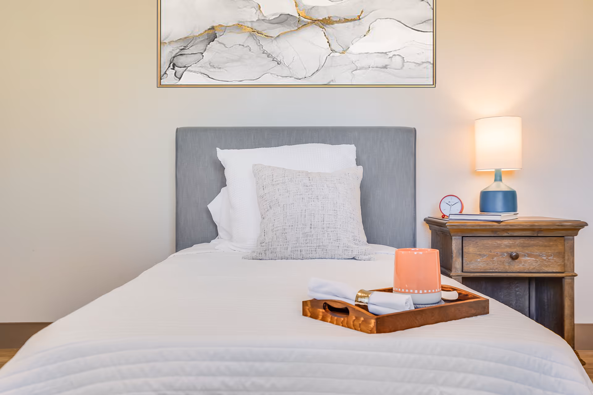 Single bed in a softly lit bedroom with a gray headboard, decorative pillows, a wooden tray with a mug, and a nightstand holding a lamp and clock.