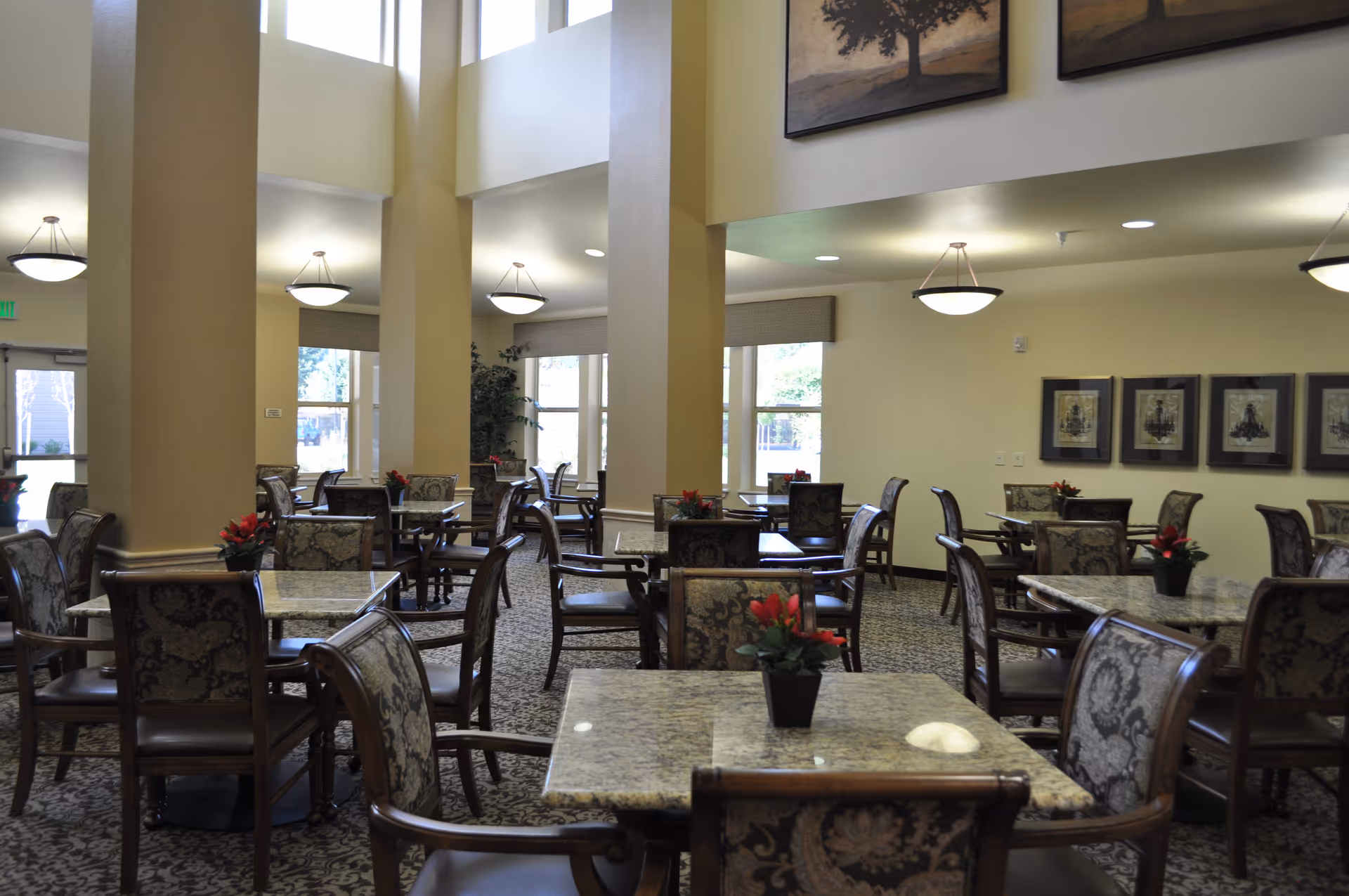 A spacious dining room with multiple square tables and upholstered chairs arranged neatly. Each table has a small potted plant with red flowers. The room features large windows allowing natural light, beige walls, patterned carpet, and framed artwork on the walls. Ceiling lights hang from above, illuminating the area.