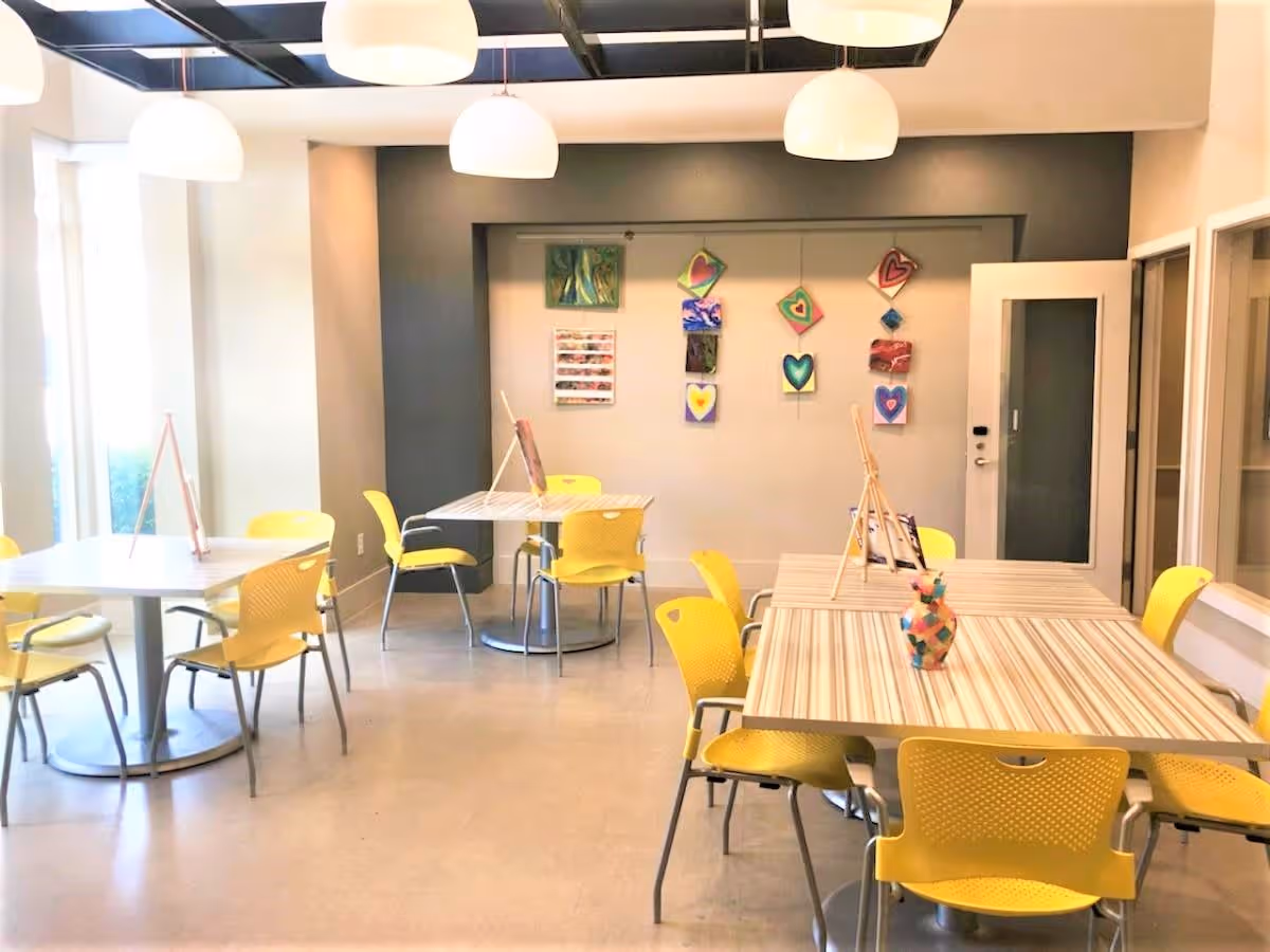 A bright activity room with several tables and yellow chairs arranged around them. The tables have easels and art supplies on them. The back wall displays colorful artwork featuring heart designs. The room is well-lit with large hanging lights and natural light from windows on the left.