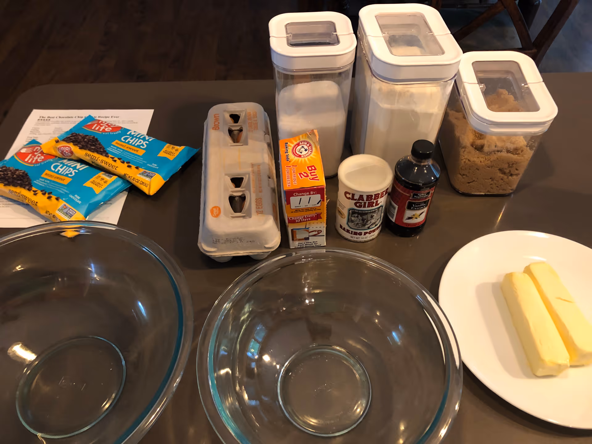 Kitchen countertop with two mixing bowls and baking ingredients laid out, including butter, eggs, flour, sugar, brown sugar, vanilla, baking powder, and chocolate chips.