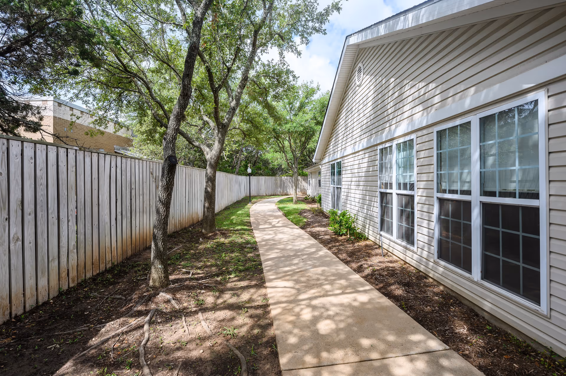 A paved walkway runs alongside a beige building with multiple windows. On the left side of the walkway, there is a wooden fence and several trees providing shade. The area has patches of grass and soil, with sunlight filtering through the tree leaves.