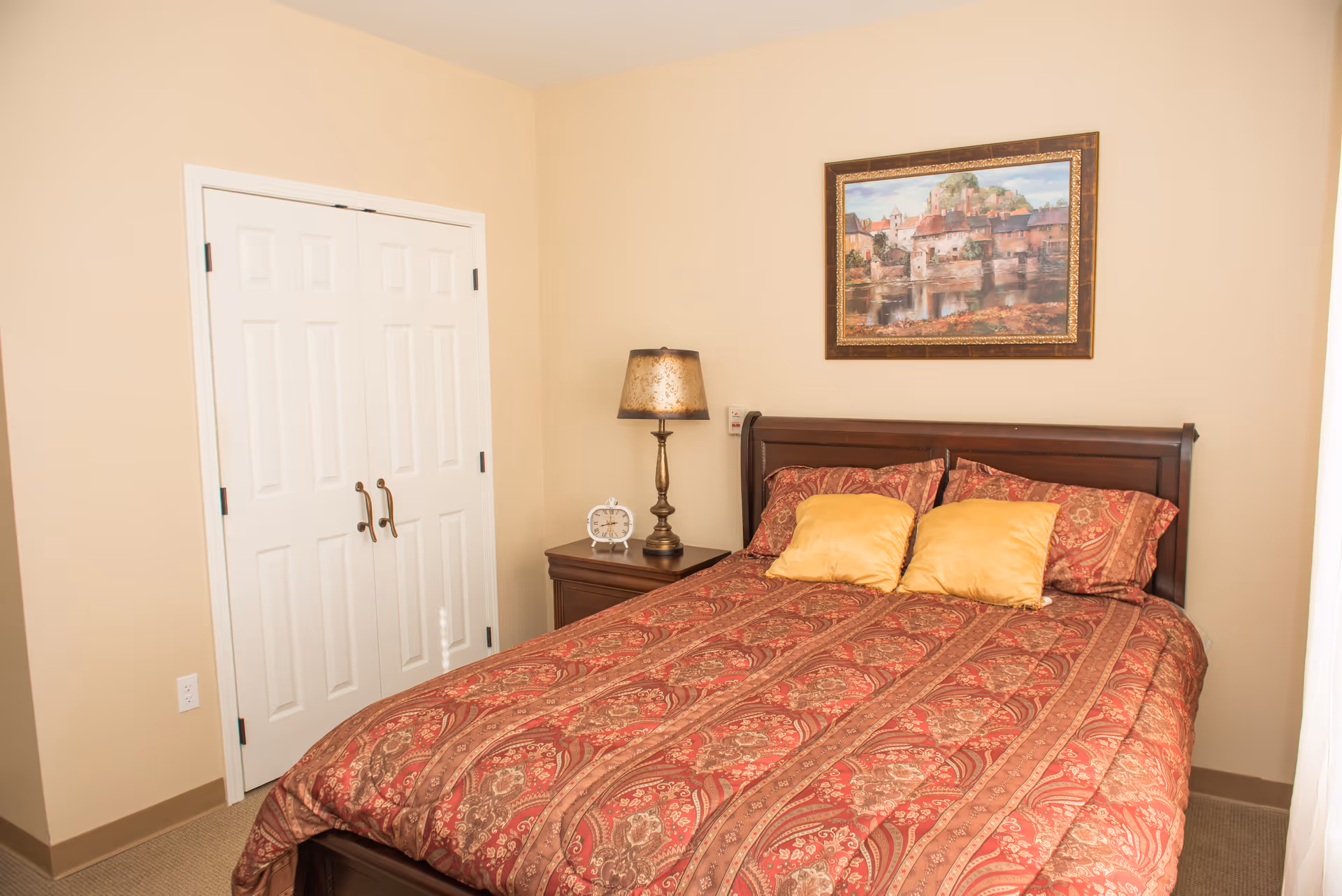 A bedroom with a wooden bed frame and a red patterned bedspread with two yellow pillows. There is a wooden nightstand with a decorative lamp and a small clock. A framed painting of a scenic village hangs on the beige wall above the bed. A white double-door closet is visible on the left side of the room.