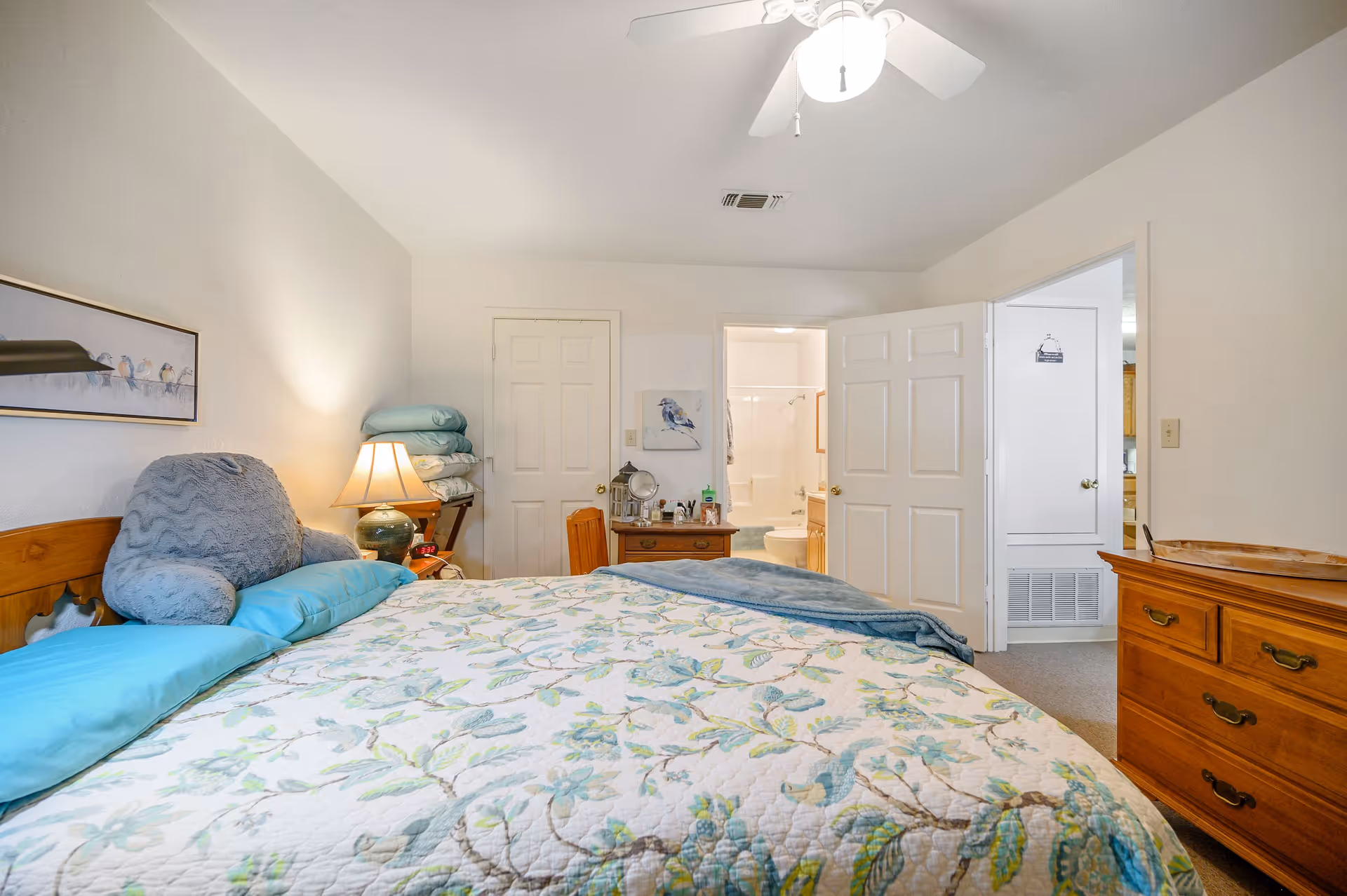A bedroom with a bed covered in a floral quilt and blue pillows. There is a wooden dresser on the right side and a nightstand with a lamp and some items on it. Two doors are visible, one leading to a bathroom with a toilet and shower, and the other leading to another room. The walls are white and there is a ceiling fan with a light.