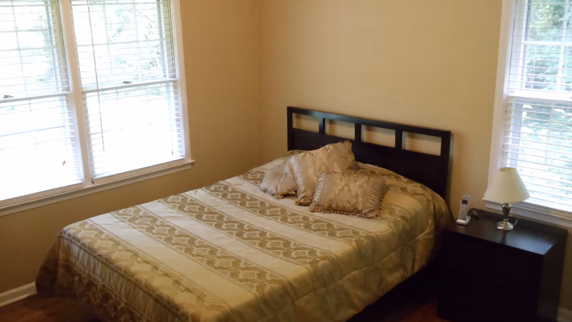A bedroom with a bed covered in a beige and gold patterned bedspread and three decorative pillows. The bed has a dark wooden headboard. There are two windows with white blinds on either side of the bed. A dark wooden nightstand with a lamp and a cordless phone is positioned next to the bed.