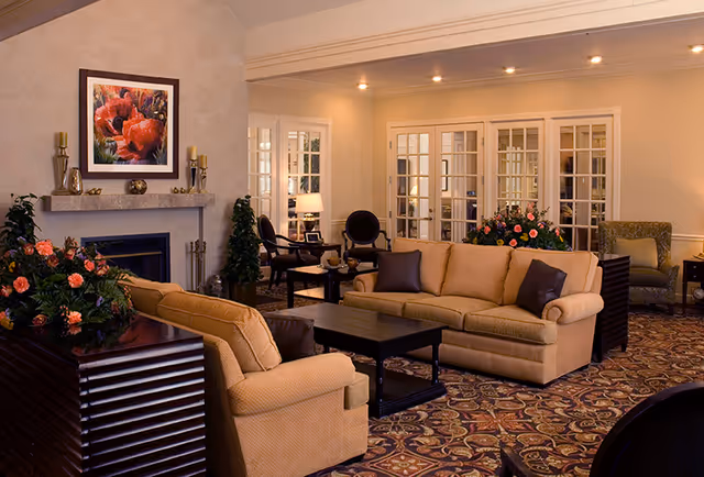 A cozy living room area with beige sofas, a dark wooden coffee table, and floral arrangements. The room features a fireplace with a framed floral painting above it, decorative candles on the mantel, and several chairs and tables in the background. The walls are light-colored, and the floor is covered with a patterned carpet. French doors with glass panes lead to another room.