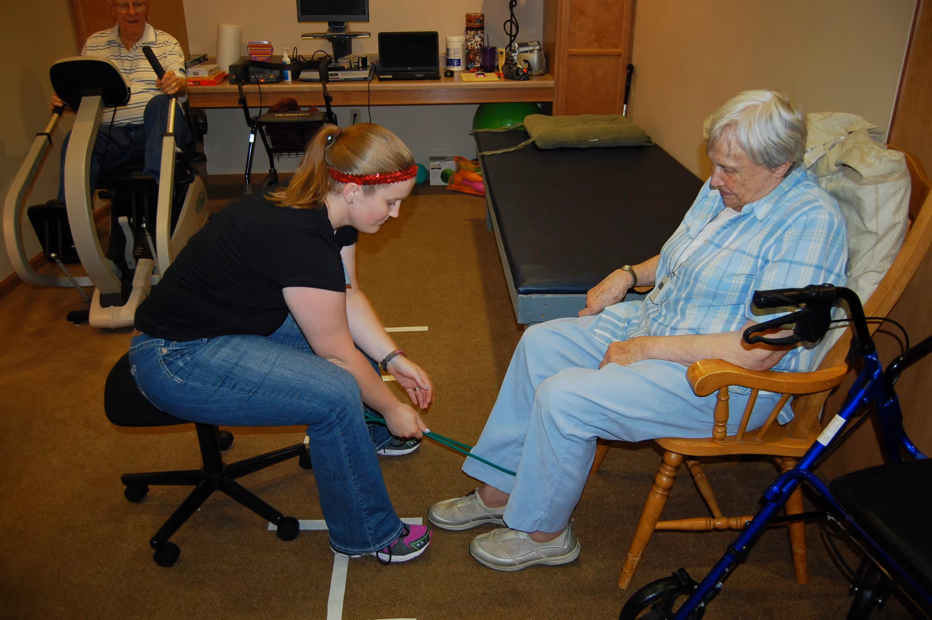 A young woman assists an elderly woman seated in a wooden chair with a resistance band exercise for her feet. In the background, an elderly man is using a stationary exercise bike in a room equipped with exercise and therapy equipment.