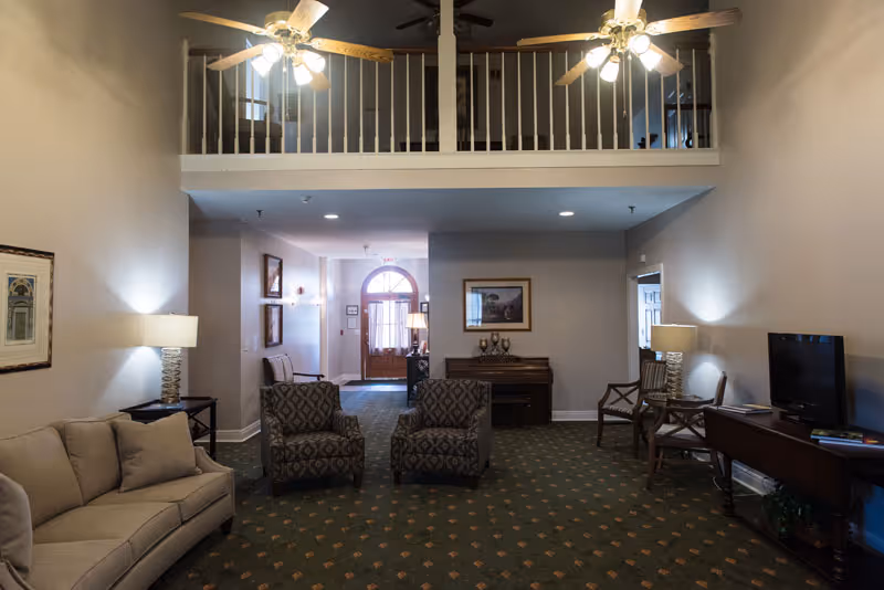 Spacious senior living community lounge with sofa, two patterned armchairs, lamps, a TV, piano, and an upstairs balcony overlooking the seating area.