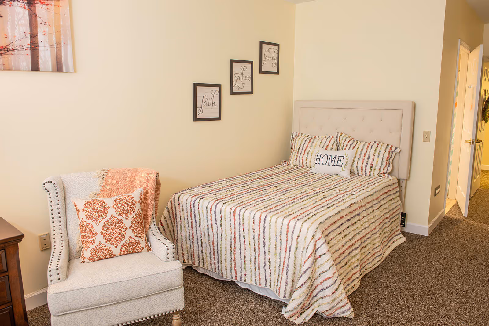 A cozy bedroom with a bed featuring a striped bedspread and pillows, including one with the word 'HOME'. Next to the bed is a cushioned armchair with a decorative pillow and a coral throw blanket. The walls are light-colored with three framed inspirational words: faith, gather, and family. A door is open, showing a hallway with carpeted flooring.