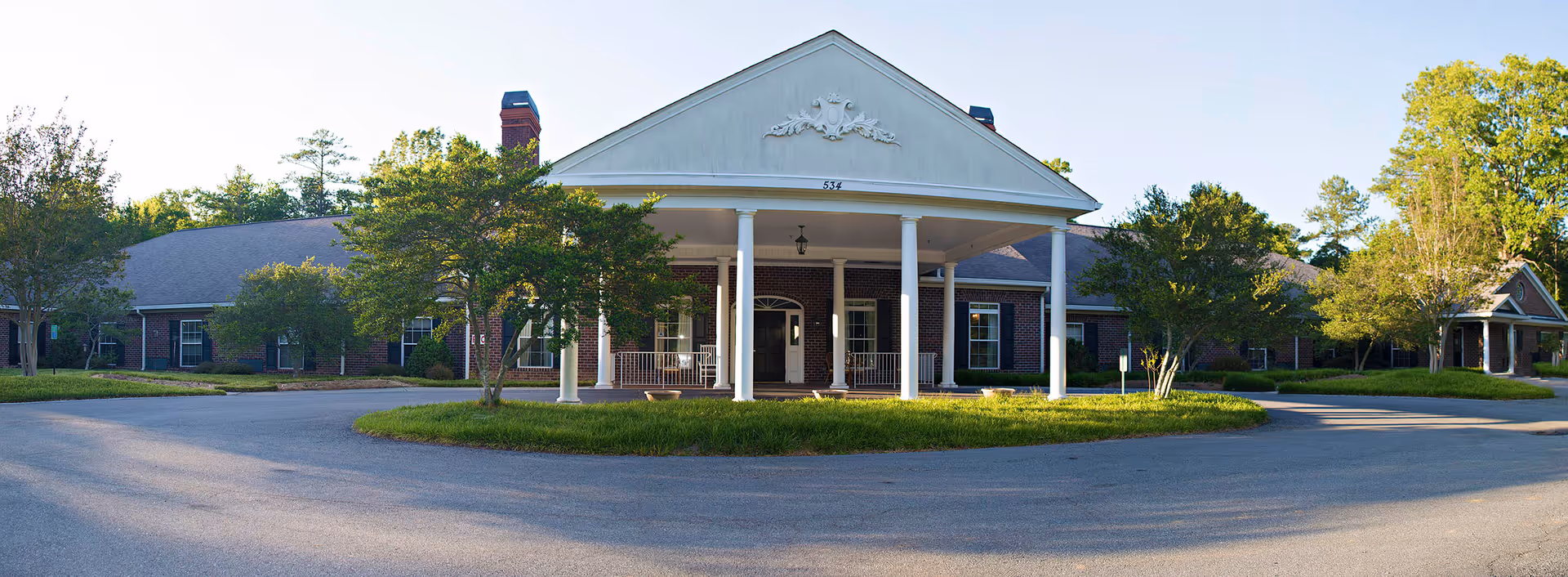 Front exterior view of Harbison Hall, a single-story brick building with white columns supporting a large triangular pediment above the entrance. The building is surrounded by trees and greenery, with a circular driveway in front.