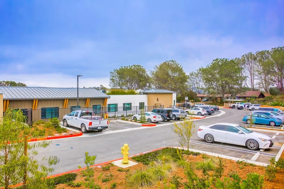 Parking lot outside a single-story building with several parked cars and a white pickup truck. The building has a metal roof and beige siding with yellow accents. There are trees and greenery surrounding the area under a partly cloudy sky.