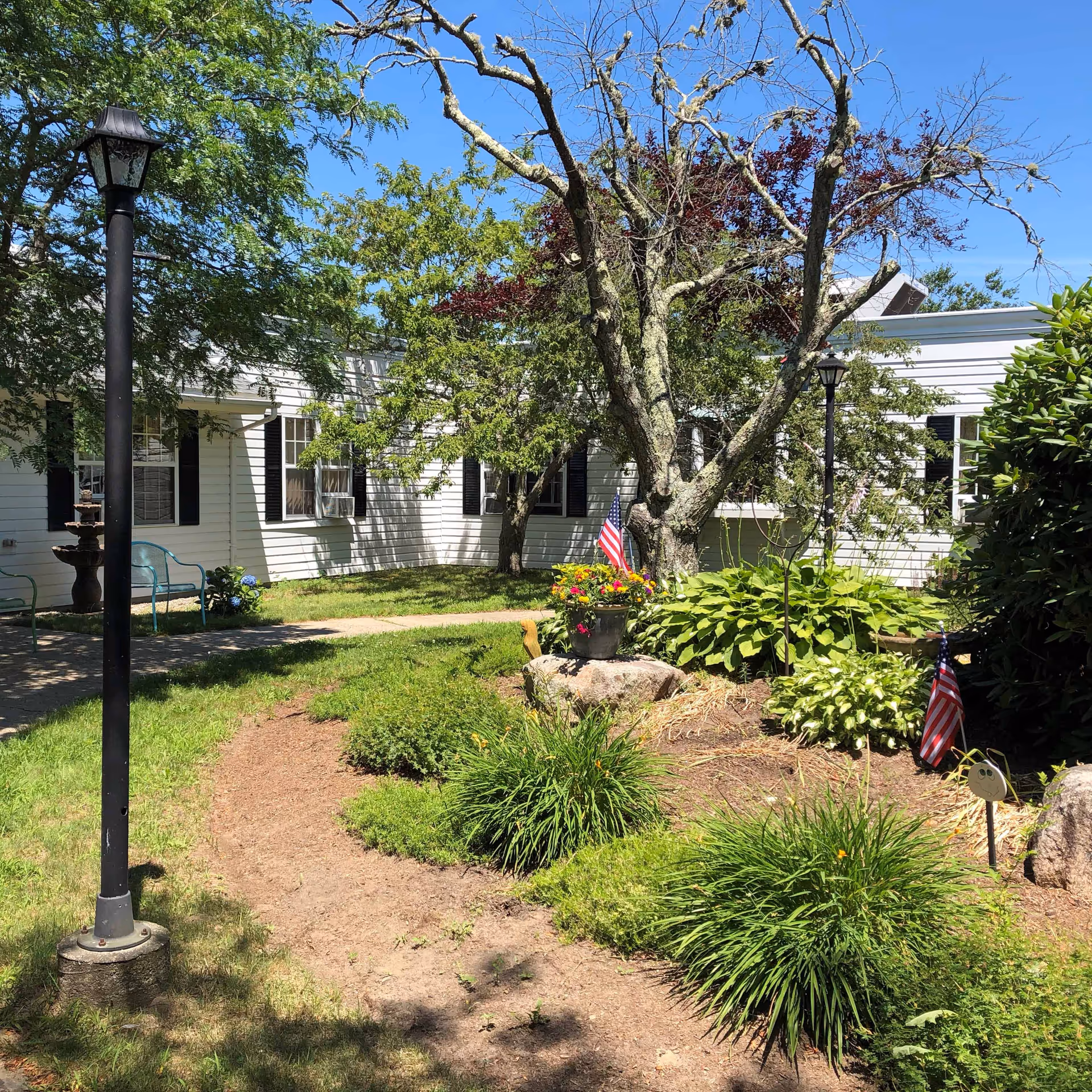 Outdoor garden area at Cape Heritage Rehabilitation & Health Care Center featuring green shrubs, a large tree with some bare branches, a flower pot with colorful flowers and an American flag, two black lamp posts, and a white building with black window shutters in the background under a clear blue sky.