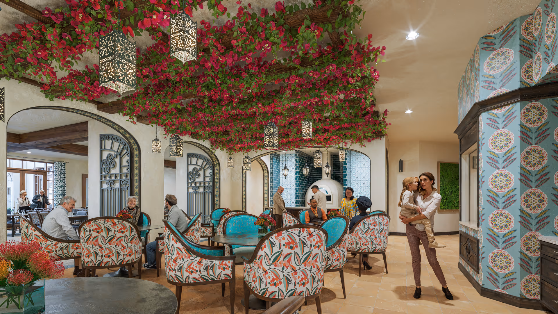 A vibrant and cozy common area in a senior living facility with floral-patterned chairs and turquoise cushions around round tables. The ceiling is decorated with hanging lanterns and lush red flowering vines. Several people are seated and standing, engaging in conversation. A woman is holding a child on the right side of the image. The walls feature decorative wrought iron and blue patterned tiles around a white oven or fireplace.