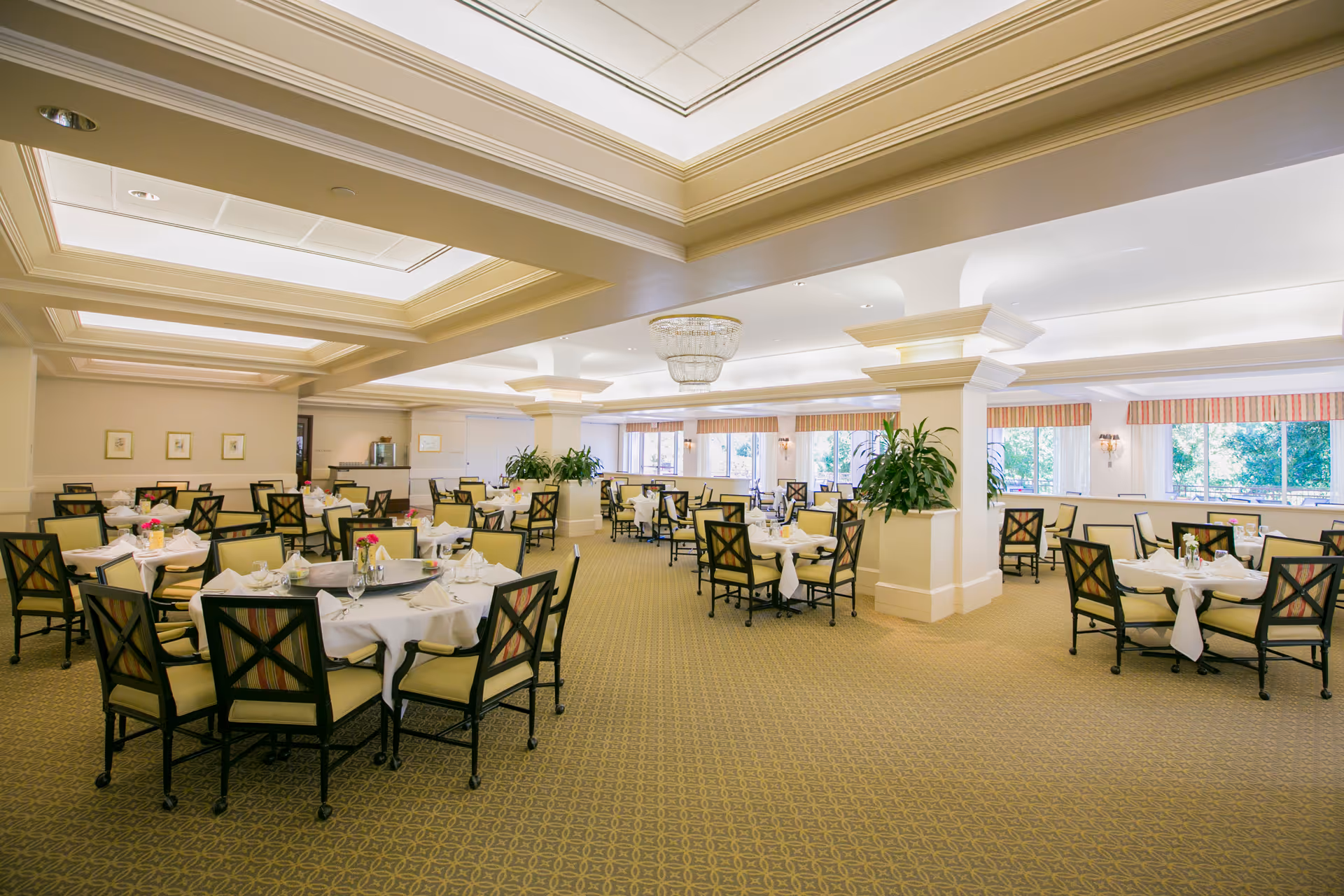 A spacious, well-lit dining room with multiple round tables covered in white tablecloths, each set with napkins, glasses, and small flower arrangements. The room features beige carpeting, large windows with striped valances, decorative columns, and a chandelier hanging from a coffered ceiling.