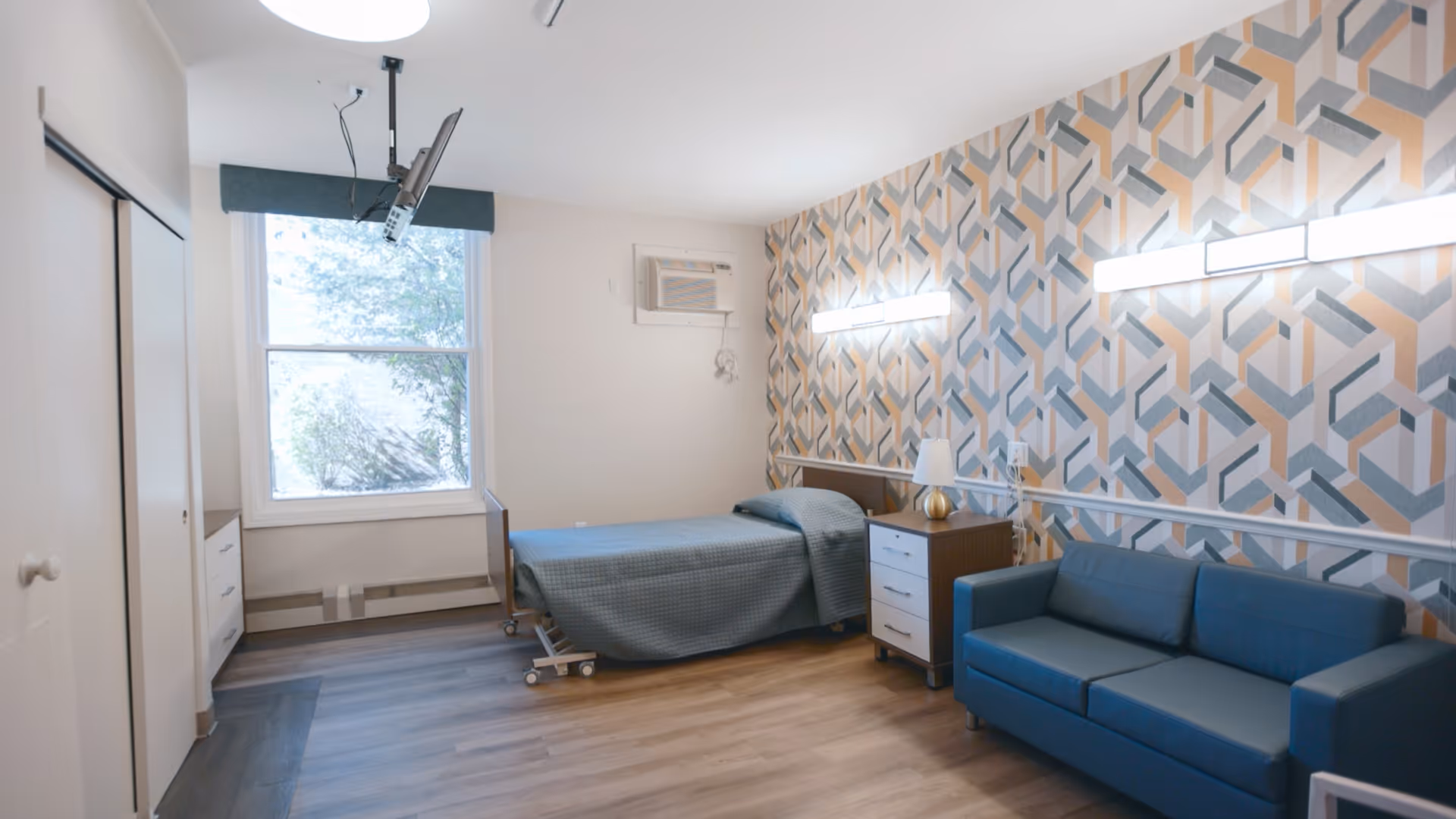 A senior living facility bedroom with a single bed covered in a blue blanket, a wooden nightstand with a lamp, a blue sofa, and a window letting in natural light. The wall behind the bed and sofa has a geometric patterned wallpaper in shades of blue, gray, and beige. The floor is wood, and there is an air conditioning unit mounted on the wall above the bed.