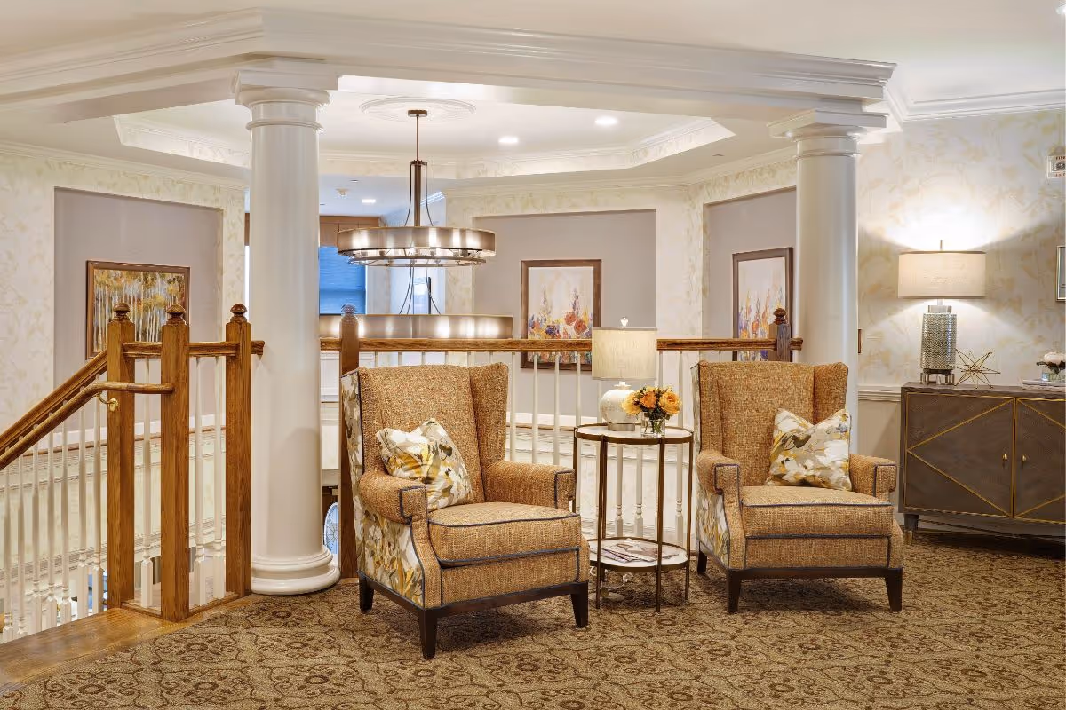 A cozy sitting area in a senior living facility featuring two upholstered armchairs with floral cushions, a small round side table with a lamp and a vase of flowers, decorative paintings on the walls, and warm lighting. The area is carpeted and has wooden railings and white columns, creating an inviting and elegant atmosphere.