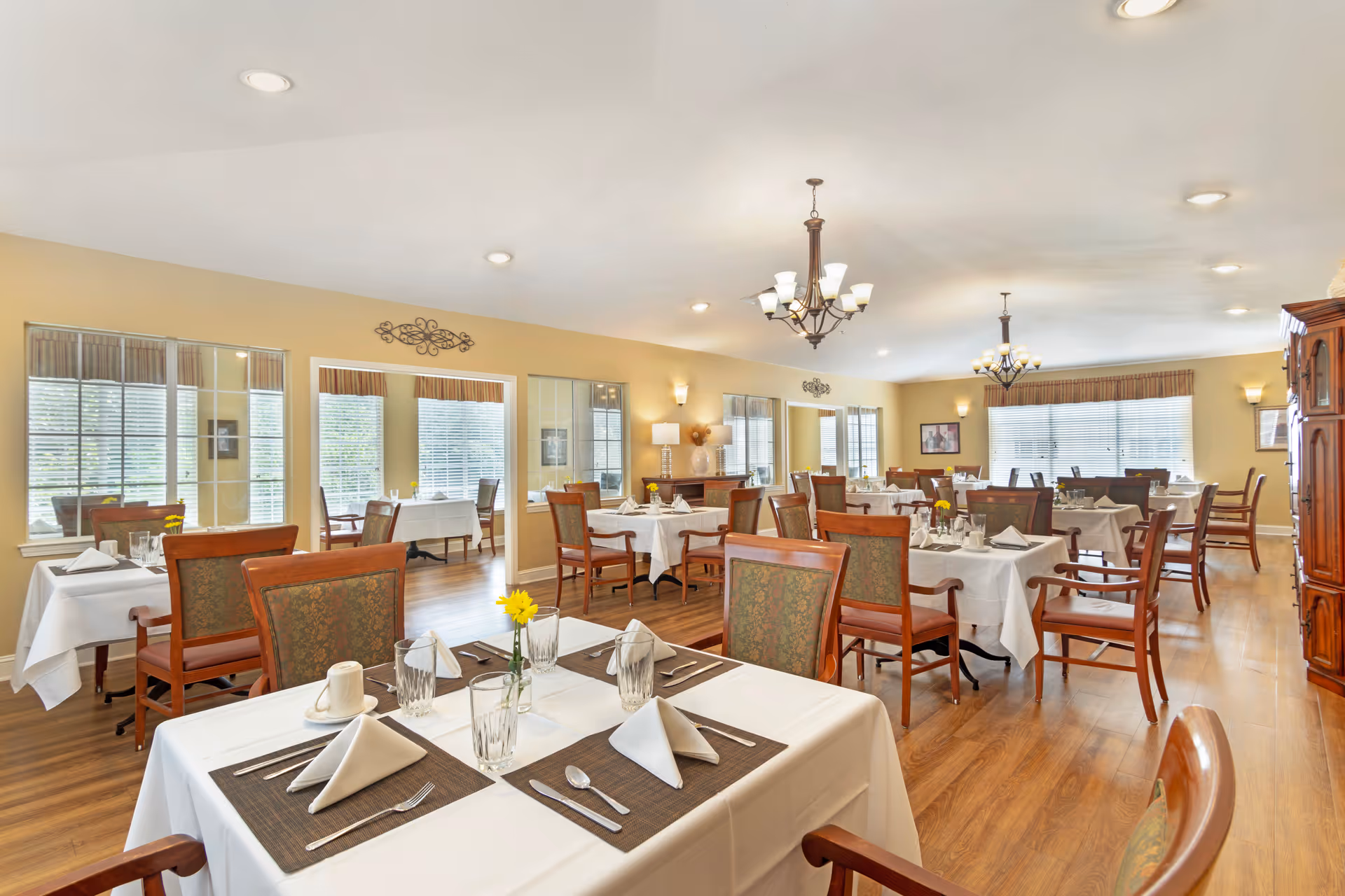 A bright and spacious dining room with multiple tables covered in white tablecloths, each set with napkins, glasses, silverware, and small yellow flower centerpieces. The room has wooden floors, large windows with blinds, and decorative light fixtures hanging from the ceiling. There are upholstered wooden chairs around the tables and a wooden cabinet on the right side.