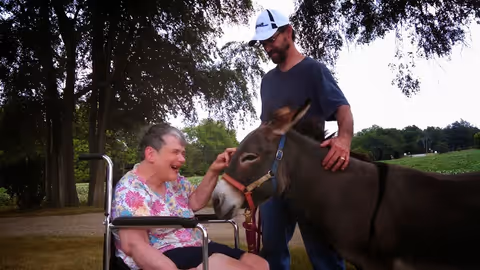 An elderly woman in a wheelchair smiling and petting a donkey while a man stands beside the donkey outdoors with trees and grass in the background.