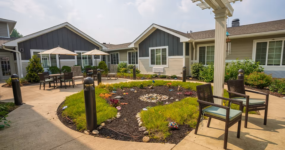 Outdoor courtyard area at Juniper Village at Hamilton featuring a circular garden with decorative stones and plants, surrounded by a concrete walkway. Several chairs and tables with umbrellas are placed around the courtyard, with a building in the background under a partly cloudy sky.
