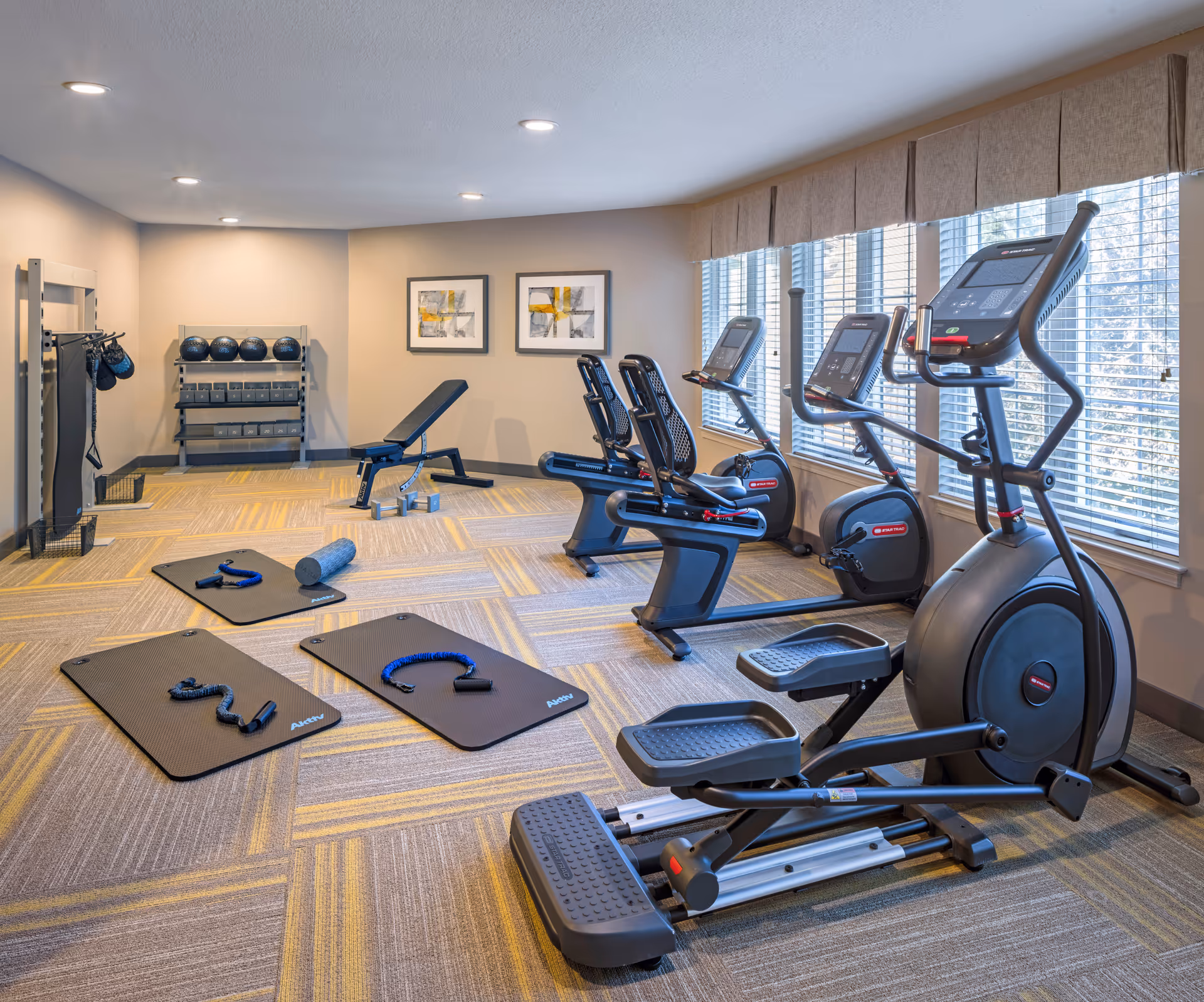 A well-lit fitness room with exercise equipment including stationary bikes, an elliptical machine, a weight bench, exercise mats with resistance bands, dumbbells, medicine balls, and wall-mounted boxing gloves. Large windows with blinds allow natural light to fill the room, and two abstract paintings hang on the far wall.