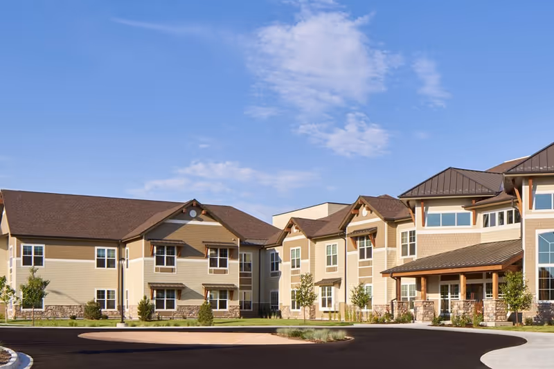 Exterior view of a large two-story assisted living facility with beige siding, stone accents, a covered entrance, and a circular driveway under a blue sky.