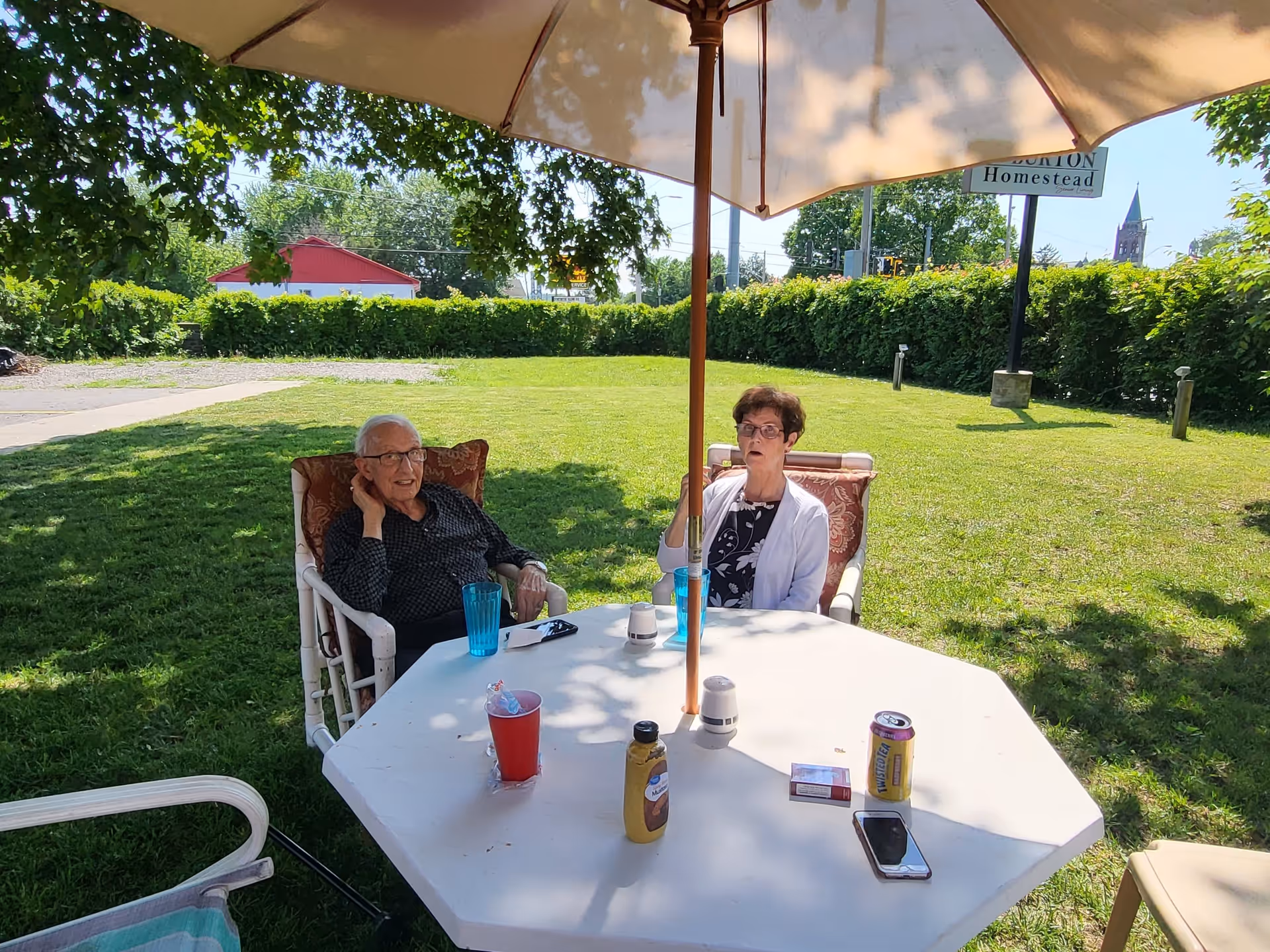 Two elderly people sitting at a white outdoor table under a large umbrella in a grassy yard. The man on the left is wearing glasses and a dark shirt, while the woman on the right is wearing glasses and a white cardigan over a patterned top. On the table are various items including a red cup, a yellow bottle, a can of Twisted Tea, a smartphone, and a pack of cigarettes. In the background, there is a green hedge, a red building, and a sign that reads 'Burton Homestead'.