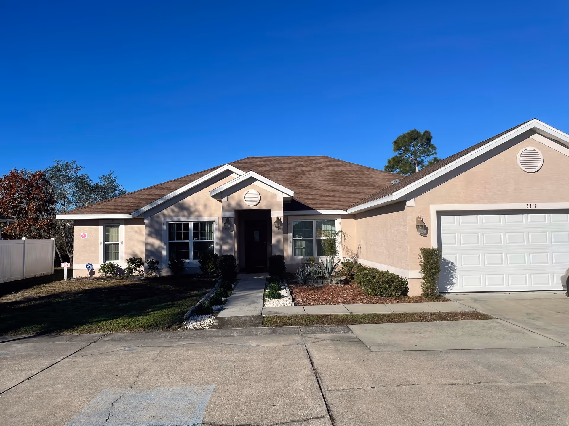 Single-story beige house with a brown shingled roof, a white garage door, and a small front garden with shrubs and a concrete walkway leading to the front door under a clear blue sky.