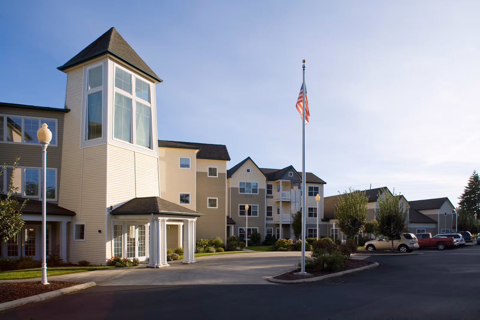 Exterior view of a multi-story senior living facility with beige siding, large windows, and a prominent tower-like entrance. There is a flagpole with an American flag in front, surrounded by landscaped greenery and a parking area with several cars.