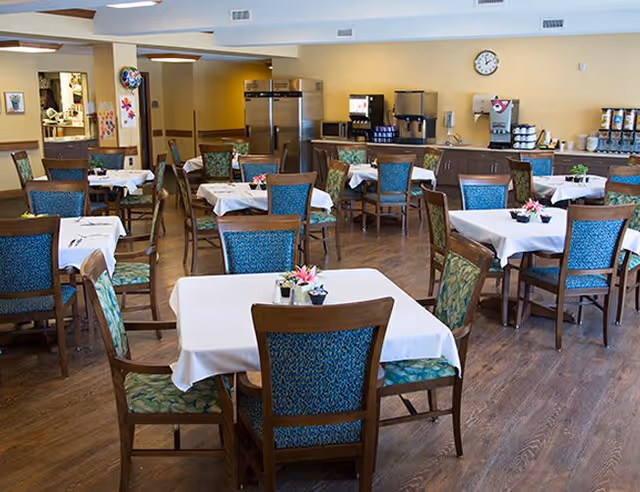 A dining room with multiple tables covered in white tablecloths, each surrounded by wooden chairs with blue and green patterned cushions. The room has a wooden floor, a clock on the wall, and a beverage station with coffee machines and dispensers in the background.