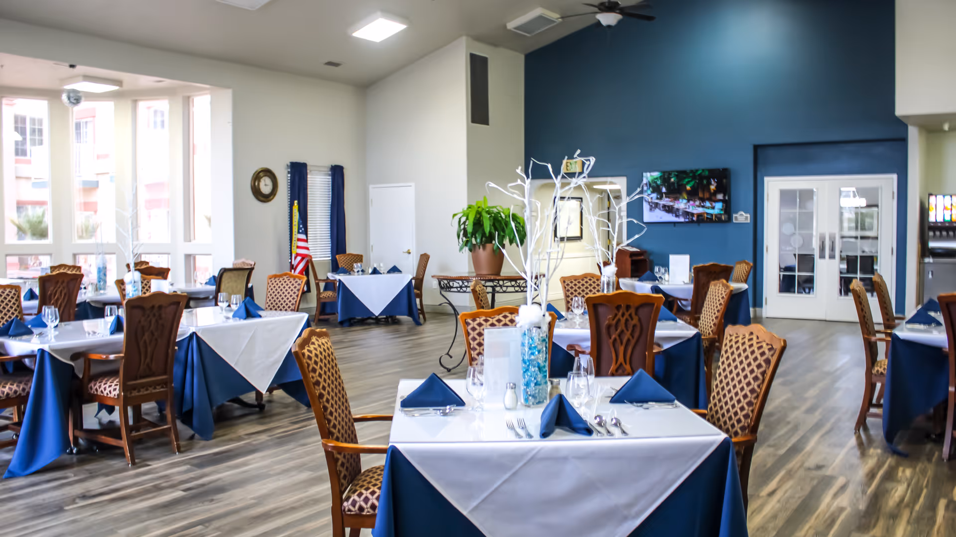 A bright dining room with multiple tables covered in white and blue tablecloths, set with glassware, silverware, and blue napkins. The room features wooden chairs with patterned upholstery, a large potted plant, a wall-mounted TV, and large windows letting in natural light. An American flag is visible near the back wall.