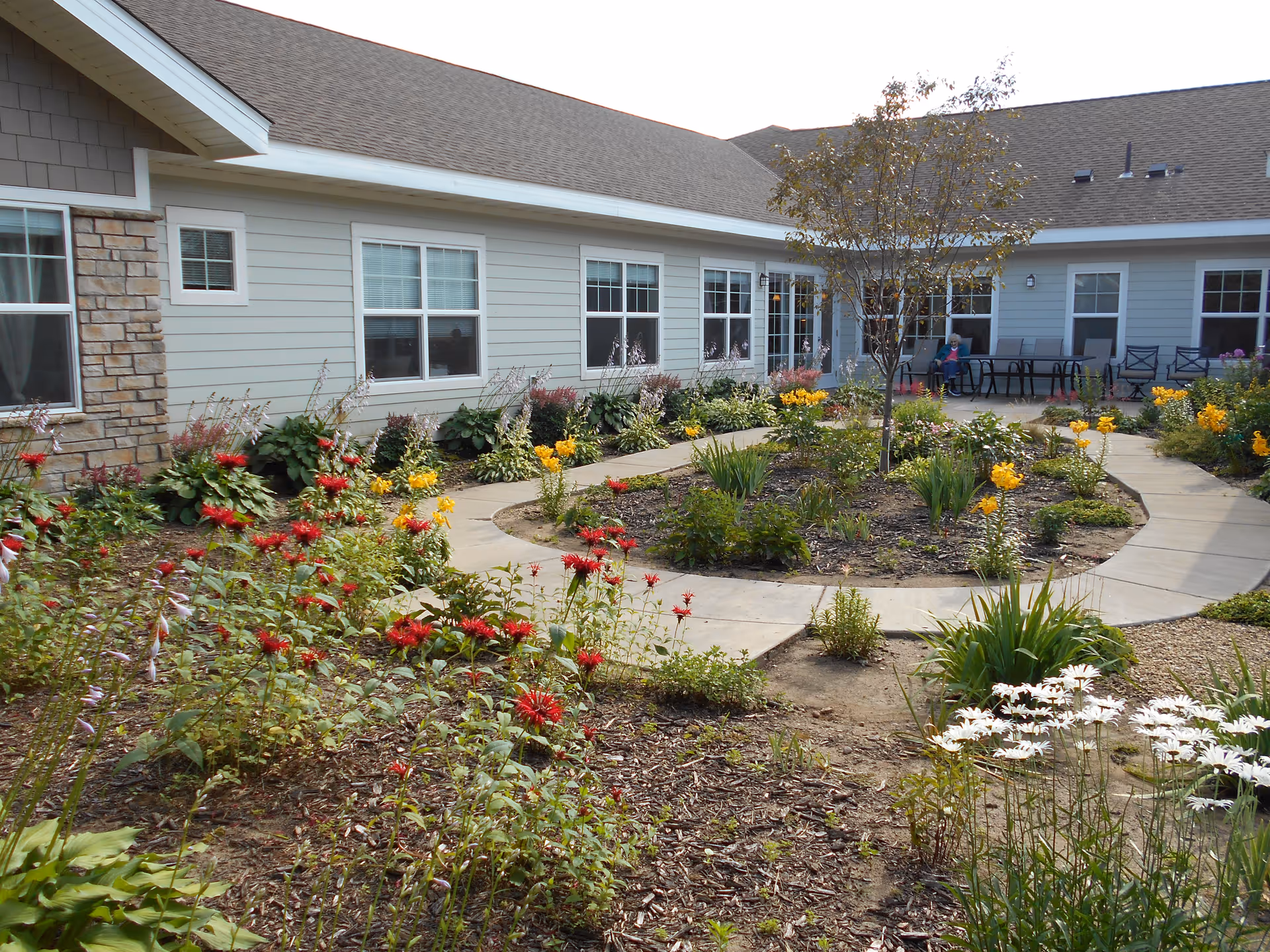 A landscaped courtyard with a circular paved walkway and colorful flower beds in front of a single-story senior living building with windows and a patio.