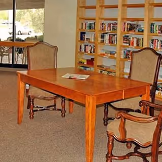 A wooden table with three upholstered chairs around it in a room with a large bookshelf filled with books. There is a glass door leading outside, and the room has carpeted flooring.