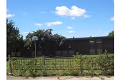 Single-story brick building with a small fenced front lawn, trees, and a blue sky with scattered clouds.