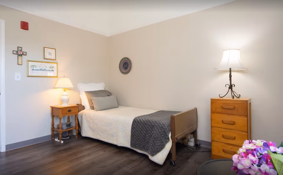 A simple, cozy bedroom in a senior living facility with a single bed covered in a light yellow patterned bedspread and a gray blanket. The bed has white and gray pillows. There is a wooden nightstand with a lamp and a clock radio to the left of the bed, and a wooden dresser with a lamp on top to the right. The walls are light beige with a few framed pictures and a decorative plate hanging. A small bouquet of purple and pink flowers is partially visible in the foreground.