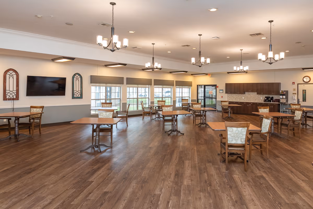Spacious dining/activity room with multiple wooden tables and chairs, chandeliers, large windows, and a kitchenette area in the back.