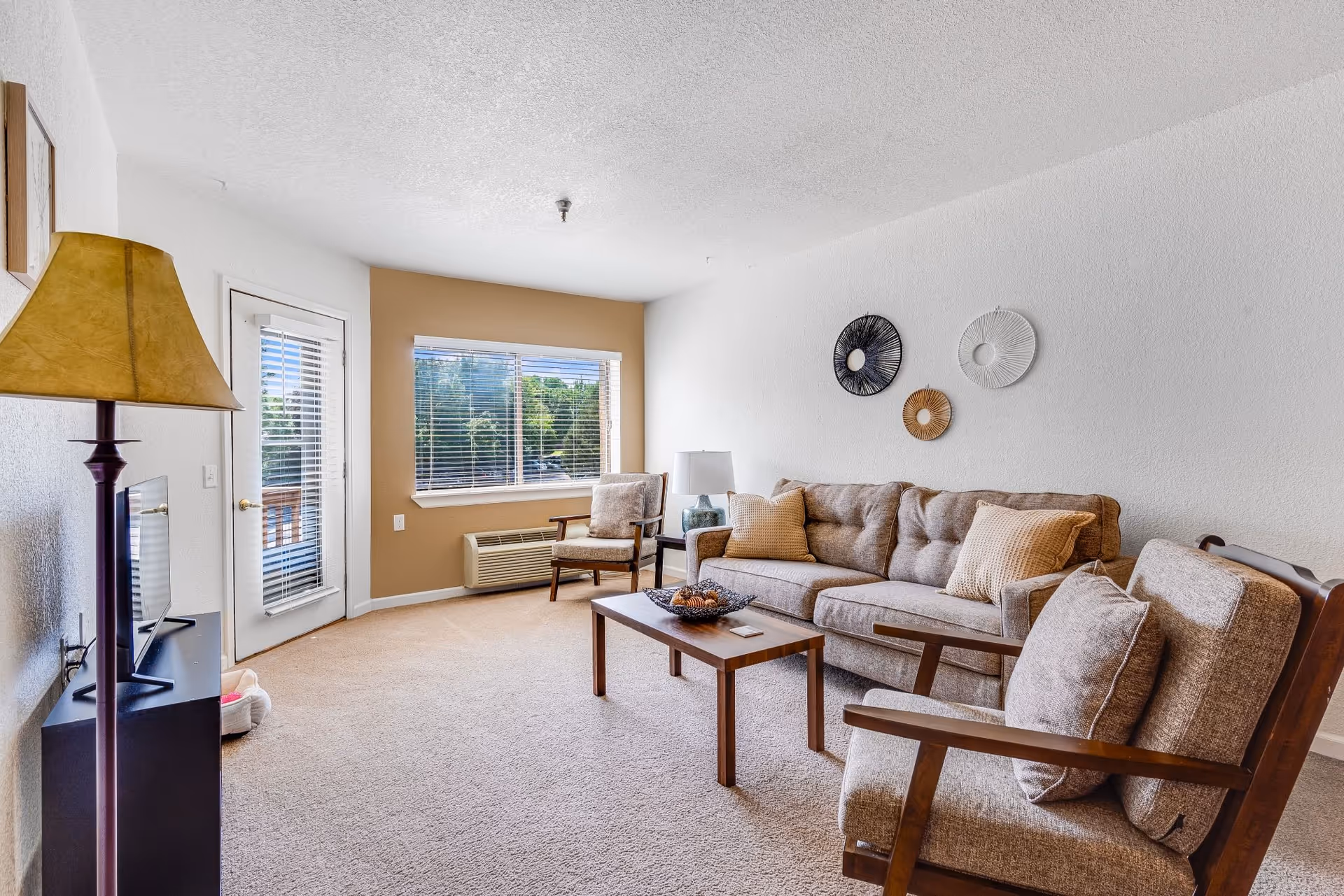 A bright living room with beige carpet, a beige sofa with cushions, two matching armchairs, a wooden coffee table with a decorative bowl, a floor lamp, a TV on a stand, and a window with blinds showing greenery outside. There is also a door leading to an outdoor area.