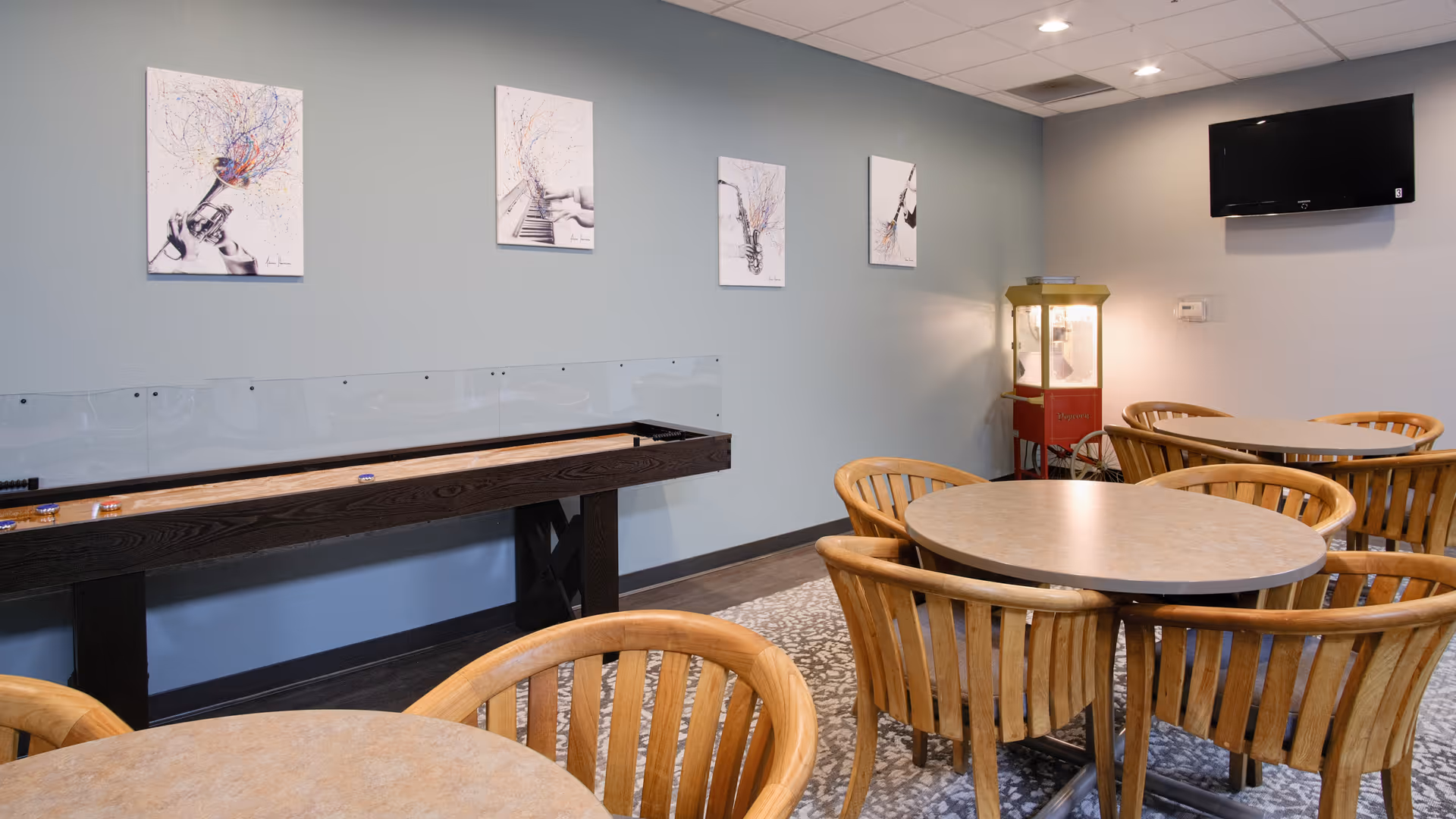 A common area with round tables and wooden chairs, a shuffleboard table along the wall, four framed musical-themed artworks hanging above the shuffleboard, a popcorn machine in the corner, and a wall-mounted television.