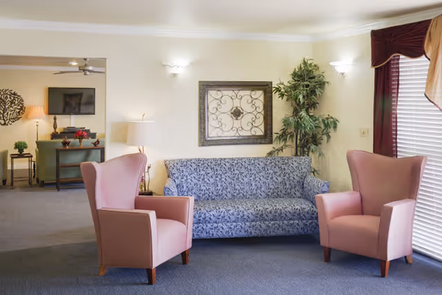 A cozy living room area with a blue patterned sofa centered against a light yellow wall, flanked by two pink armchairs. A decorative metal wall art piece hangs above the sofa, and a tall green plant is placed in the corner. To the left, there is a glimpse of another room with a TV mounted on the wall, a green chair, and a side table with a lamp and decorative items. Window blinds with burgundy valances are on the right side, allowing natural light to enter.