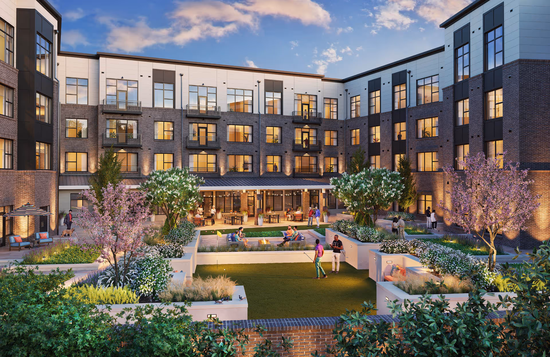 Courtyard of a multi-story senior living building with landscaped lawns, seating areas, and people socializing at dusk.